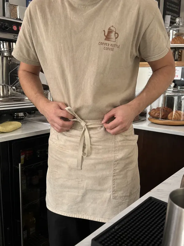 A barista tying an apron over a sand Copper Kettle Coffee shirt